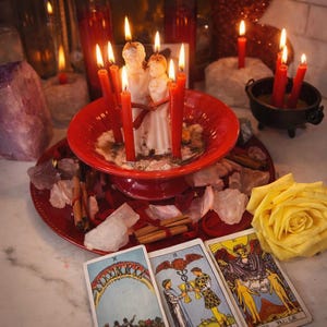 May include: A spiritual arrangement featuring lit red candles, crystals, tarot cards, and a yellow rose. A central red dish holds a white candle figure surrounded by red candles. Cinnamon sticks and crystals are scattered around the base.