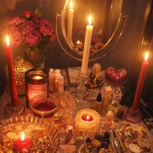 May include: A candlelit display featuring a mirror, red roses, and various items. Lit candles of different sizes and shapes are arranged on a table, along with small bottles, crystals, and a heart-shaped candle. The scene is bathed in warm light.