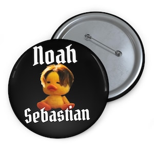 May include: A black button pin featuring a plush duck with brown hair and an orange beak. The text "Noah Sebastian" is printed in white gothic lettering. The pin has a silver metal backing with a pin.