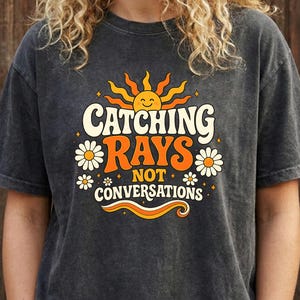 May include: Dark gray t-shirt with the text "Catching Rays Not Conversations" in a retro design. The graphic includes a smiling sun, daisies, and orange accents. The shirt is a casual wear item.