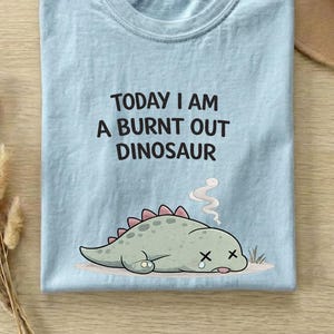Cute sad dinosaur shirt with burnout theme displayed on chambray t-shirt in neutral Etsy style mockup