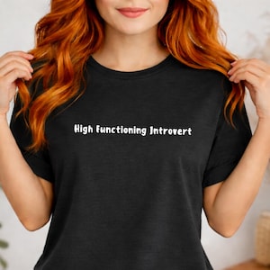 Könnte beinhalten: Schwarzes T-Shirt mit dem weißen Text "High functioning Introvert". Das Shirt hat einen Rundhalsausschnitt und scheint aus einem weichen Material zu sein. Das Model trägt eine blaue Jeans.
