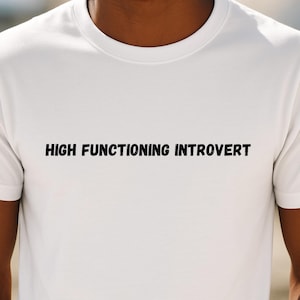 Könnte beinhalten: Weißes T-Shirt mit dem Text "HIGH FUNCTIONING INTROVERT" in Schwarz auf der Brust gedruckt. Das T-Shirt hat einen Rundhalsausschnitt und kurze Ärmel. Das Bild ist eine Nahaufnahme des T-Shirts.