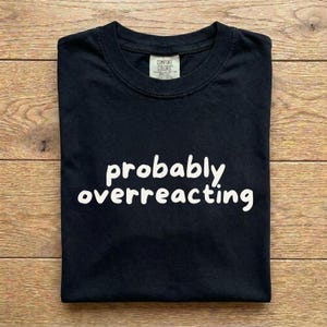 May include: A black short-sleeved t-shirt with a crew neckline. The centre of the chest features the white text "probably overreacting" in a rounded, casual font. A "Comfort Colors" brand label is visible on the inside of the collar.