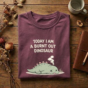 Burnt out dinosaur shirt on Comfort Colors berry t-shirt with cute tired dino graphic and funny burnout quote
Cute burnout dinosaur graphic tee shown on folded Comfort Colors shirt with soft aesthetic styling and rustic background
