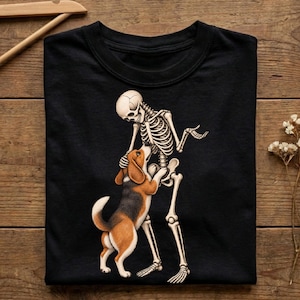 May include: A black crew neck t-shirt featuring a graphic illustration of a human skeleton standing upright and interacting with a brown, black, and white beagle dog. The skeleton is reaching out to pet the dog, which is standing on its hind legs.