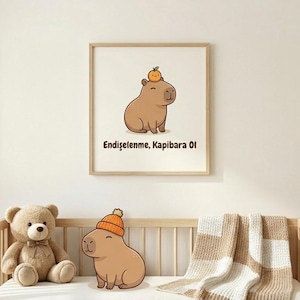 Pode incluir: Uma impressão emoldurada com um capivara de desenho animado com uma pequena fruta laranja na cabeça e o texto "Endişelenme, Kapibara Ol". Um urso de pelúcia e um bicho de pelúcia capivara com um chapéu estão em um berço de madeira.
