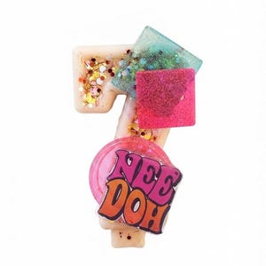 May include: A cream-colored number seven shaped wax melt embedded with multicolored glitter. It features a translucent teal square, a solid pink square, and a circular pink topper with the text NEE DOH printed in bold purple and orange letters.