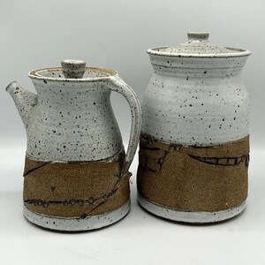 May include: A ceramic pitcher and jar set. The pitcher has a spout and handle, and the jar has a lid. Both are speckled white with a brown band around the base. The set is handmade and has a rustic aesthetic.