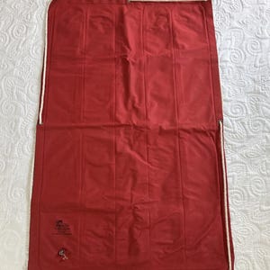 Puede incluir: Una bolsa rectangular de lona roja con ribete de cuerda blanca y una pequeña válvula metálica. La bolsa tiene costuras verticales y está colocada plana sobre una superficie blanca. Se ve un pequeño logotipo impreso.