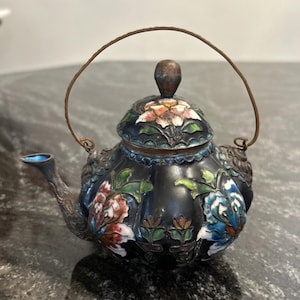 May include: A small, ornate teapot with a black base and floral designs in red, white, and blue. The teapot has a metal handle and spout, and a lid with a decorative knob. The teapot is likely made of metal and enamel.