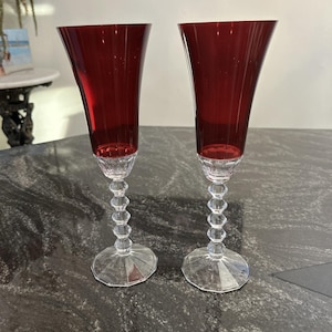 Könnte beinhalten: Zwei elegante rote Sektgläser mit klaren, facettierten Stielen und Böden. Die rubinroten Kelche verjüngen sich nach oben und erzeugen eine elegante Silhouette. Die Stiele weisen eine Reihe klarer, runder, dekorativer Elemente auf.