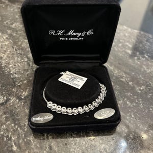 May include: A sterling silver bracelet with gemstones, displayed in a black jewelry box from R.H. Macy & Co. The box is open, revealing the bracelet's design. A tag with details is attached.