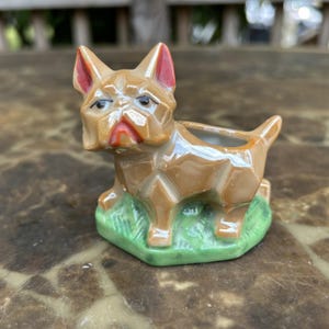 Pode incluir: Estatueta de cão em cerâmica castanha com detalhes vermelhos nas orelhas e na boca. O cão está posicionado sobre uma base verde, com uma pequena abertura nas costas. A estatueta tem um design geométrico.