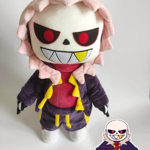 Op de afbeelding: Een pluche pop van een personage met een wit gezicht, zwarte ogen met rode pupillen en een brede, grijnzende glimlach. De pop draagt een paarse jas, korte broek en een rood shirt, met gele accenten. Een kleinere sticker van het personage is ook aanwezig.
