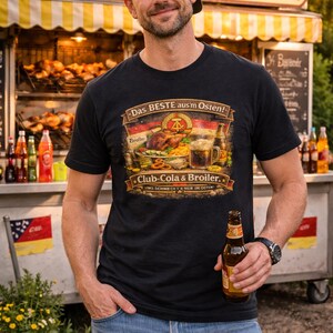May include: Black t-shirt with the text "Das BESTE aus'm Osten!" and a graphic of food and drinks. The person is holding a beer bottle. The background includes a food stand with the word "BROER" on top.
