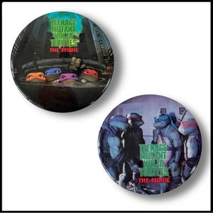 May include: Two Teenage Mutant Ninja Turtles movie pinback buttons. The first button features the four turtles in a sewer scene with the text "Teenage Mutant Ninja Turtles The Movie". The second button features the four turtles in a more modern setting with the text "Teenage Mutant Ninja Turtles The Movie".