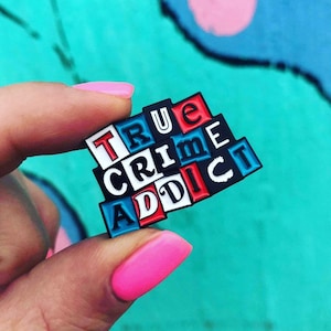May include: A colorful enamel pin with the words "TRUE CRIME ADDICT" in black letters on a white background. The letters are arranged in a staggered pattern, with each letter on a separate square. The squares are red, white, and blue.