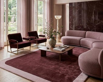 Op de afbeelding: Een woonkamerset met een gebogen roze bank met textuur, twee donkerbordeauxrode fluwelen fauteuils met gouden frames, een houten salontafel, een groot tweekleurig bordeaux en roze vloerkleed en een hoge stenen vloerlamp.