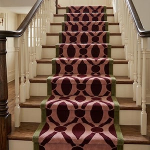 May include: A staircase featuring a carpet runner with a repeating geometric pattern in dark burgundy and light pink, bordered by a solid olive green trim. The stairs have dark wood treads and white risers, flanked by white balusters and dark wood handrails.