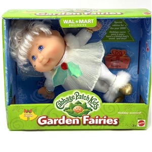 Puede incluir: Una muñeca Cabbage Patch Kids Garden Fairies en una caja verde. La muñeca tiene cabello blanco, ojos azules y viste un vestido blanco con un acento de hoja de acebo verde. El empaque incluye texto como Wal-Mart Exclusive y Golden Holiday.