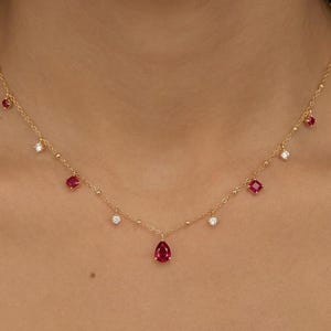Könnte beinhalten: Eine zarte Goldkette mit kleinen, runden Goldperlen und abwechselnd roten und klaren Edelsteinen. Die Halskette hat einen tropfenförmigen roten Edelsteinanhänger in der Mitte, mit quadratischen und runden roten Edelsteinen und klaren Edelsteinen.