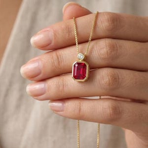 May include: A gold necklace featuring a rectangular emerald-cut red gemstone pendant in a bezel setting, topped with a small round white diamond accent. The pendant hangs from a delicate gold box chain, showcasing a polished metal finish.