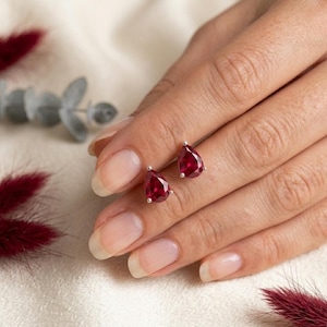 May include: A pair of stud earrings featuring pear-shaped, deep red gemstones. The earrings are set in silver-colored metal and are displayed on a hand. The gemstones have a vibrant, polished appearance, reflecting light.