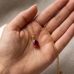 May include: A gold-colored necklace featuring a teardrop-shaped, deep red gemstone pendant. The pendant is suspended from a delicate chain. The necklace is displayed against a neutral background.