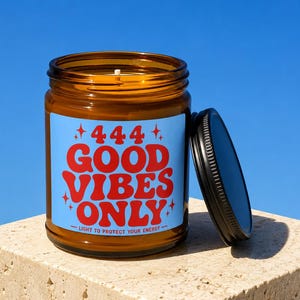 Könnte beinhalten: Ein braunes Kerzenglas mit einem hellblauen Etikett, das den roten Schriftzug 444 GOOD VIBES ONLY und LIGHT TO PROTECT YOUR ENERGY sowie kleine rote Sterne zeigt. Das Glas hat einen schwarzen Metalldeckel, der daneben platziert ist.