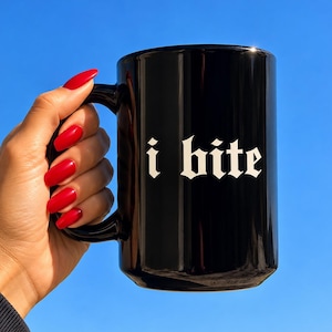 Puede incluir: Una taza de cerámica negra brillante con el texto i bite impreso en una fuente gótica blanca. La taza tiene un asa curva y un cuerpo cilíndrico, mostrando una superficie lisa y reflectante sobre un fondo de cielo azul claro.