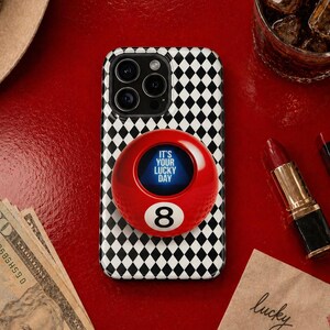 May include: A red and black phone case with a black and white diamond pattern, featuring a red 8-ball phone grip. The 8-ball grip has the text "IT'S YOUR LUCKY DAY" and the number 8. Other items include a lipstick, cash, and a drink.