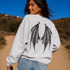 May include: A light grey crewneck sweatshirt featuring a large, detailed black graphic of bat-like wings printed on the back. The sweatshirt has long sleeves and a relaxed fit, paired with light blue denim shorts with a frayed hemline.