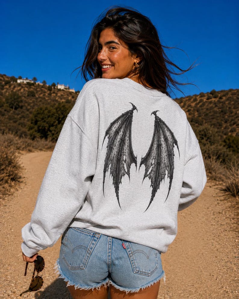 May include: A light grey crewneck sweatshirt featuring a large, detailed black graphic of bat-like wings printed on the back. The sweatshirt has long sleeves and a relaxed fit, paired with light blue denim shorts with a frayed hemline.