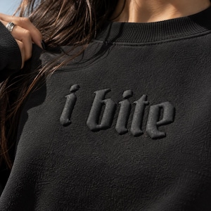 May include: A black long-sleeve sweatshirt featuring the text i bite in a raised, embossed, gothic-style font across the chest. The garment has a ribbed crew neckline and a textured fabric finish, presenting a monochromatic aesthetic throughout.