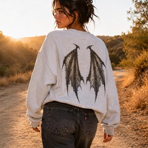 May include: A light grey crewneck sweatshirt featuring a large, detailed black graphic of bat-like wings on the back, paired with dark charcoal grey denim jeans. The sweatshirt has long sleeves and a relaxed fit, showcasing a dark, intricate wing design.