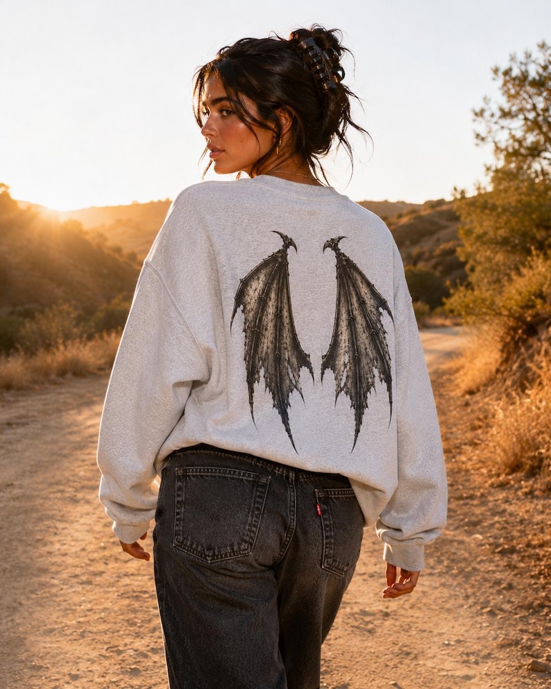 May include: A light grey crewneck sweatshirt featuring a large, detailed black graphic of bat-like wings on the back, paired with dark charcoal grey denim jeans. The sweatshirt has long sleeves and a relaxed fit, showcasing a dark, intricate wing design.