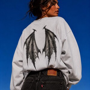 May include: A light grey crewneck sweatshirt featuring a large, detailed black graphic of bat-like wings printed on the back. The sweatshirt is paired with black denim jeans that have a brown leather patch on the waistband and a small red tag.