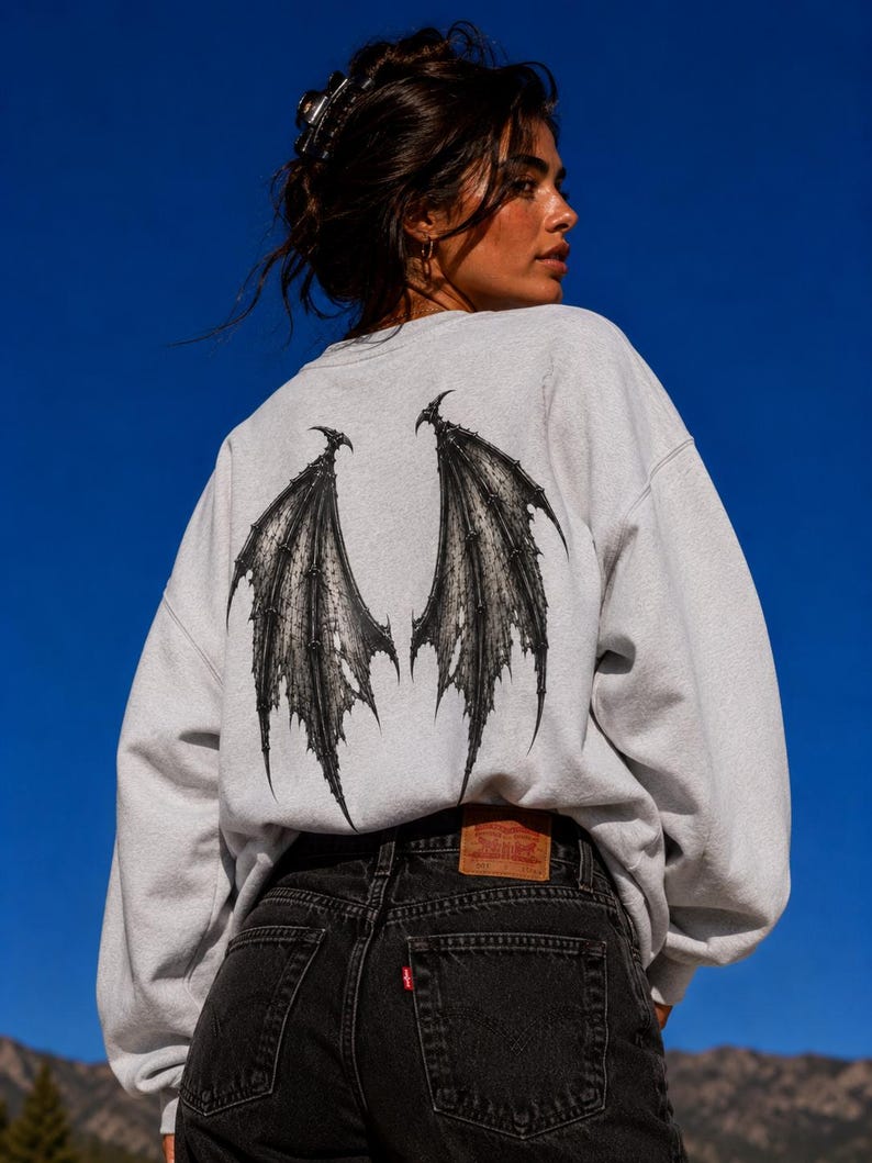 May include: A light grey crewneck sweatshirt featuring a large, detailed black graphic of bat-like wings printed on the back. The sweatshirt is paired with black denim jeans that have a brown leather patch on the waistband and a small red tag.