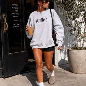 May include: A light grey crewneck sweatshirt featuring the word devilish printed in a black gothic-style font across the chest. The long-sleeved pullover has a ribbed neckline, cuffs, and hem, providing a relaxed fit for casual daily wear.