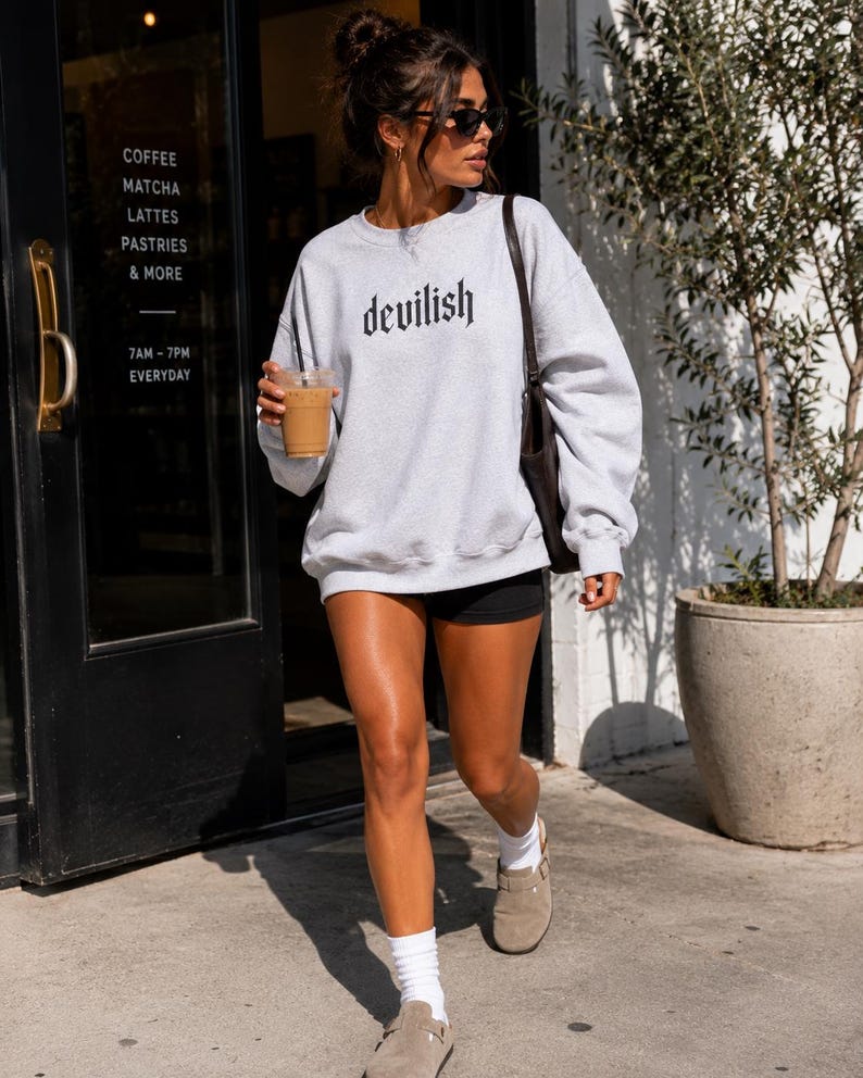 May include: A light grey crewneck sweatshirt featuring the word devilish printed in a black gothic-style font across the chest. The long-sleeved pullover has a ribbed neckline, cuffs, and hem, providing a relaxed fit for casual daily wear.
