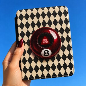 Puede incluir: Una funda rectangular para tableta con un patrón de rombos arlequín en negro y crema. Centrado en la funda hay un accesorio circular rojo translúcido que recuerda a una bola ocho, con un triángulo rojo y el texto blanco LOVE SUCKS.