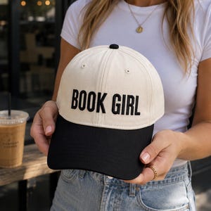 May include: A cream-colored baseball cap with a black brim and the embroidered text "BOOK GIRL" in bold, black letters. The cap has a black button on top and is a classic six-panel design.