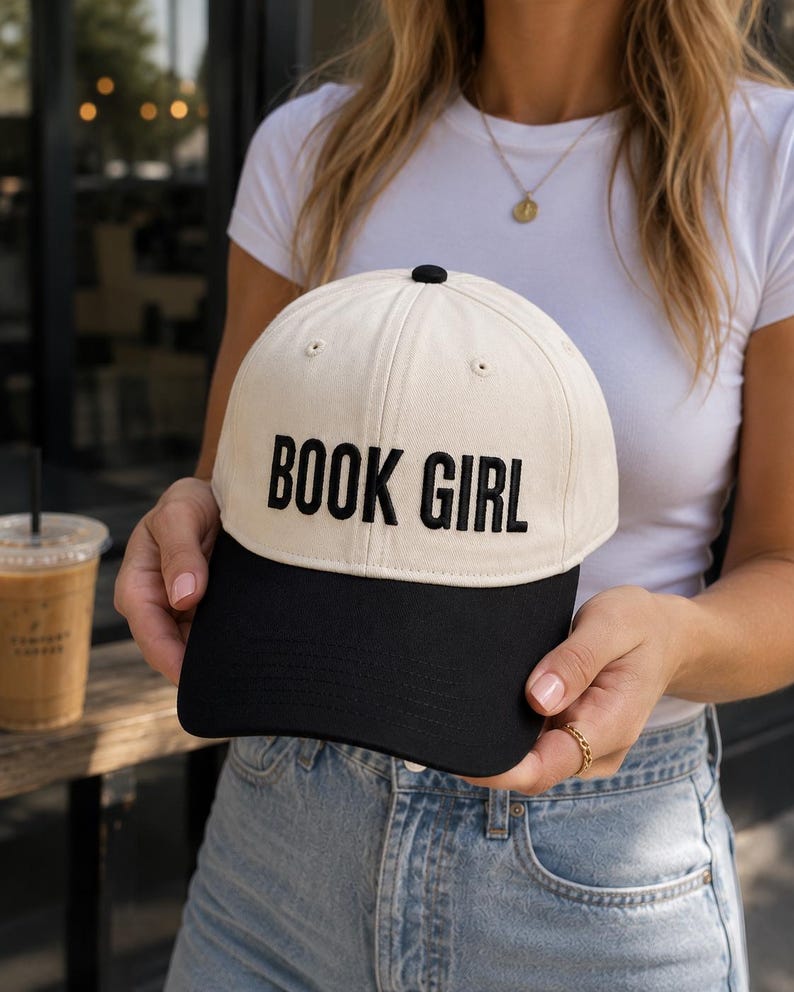 May include: A cream-colored baseball cap with a black brim and the embroidered text "BOOK GIRL" in bold, black letters. The cap has a black button on top and is a classic six-panel design.