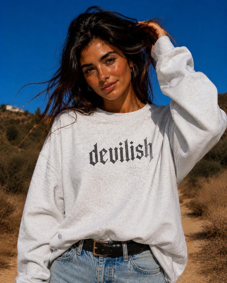 May include: A light grey crewneck sweatshirt featuring the word devilish printed in a black gothic-style font across the chest. The garment has long sleeves with ribbed cuffs and a ribbed neckline, paired with blue denim jeans and a black belt.