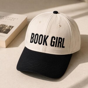 May include: A cream-colored baseball cap with a black brim and the embroidered text "BOOK GIRL" in bold, black letters. A book with the title "THE SECRET HISTORY" is visible in the background.