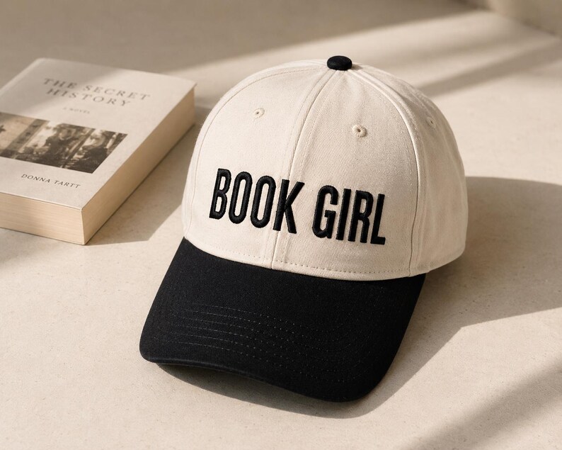 May include: A cream-colored baseball cap with a black brim and the embroidered text "BOOK GIRL" in bold, black letters. A book with the title "THE SECRET HISTORY" is visible in the background.