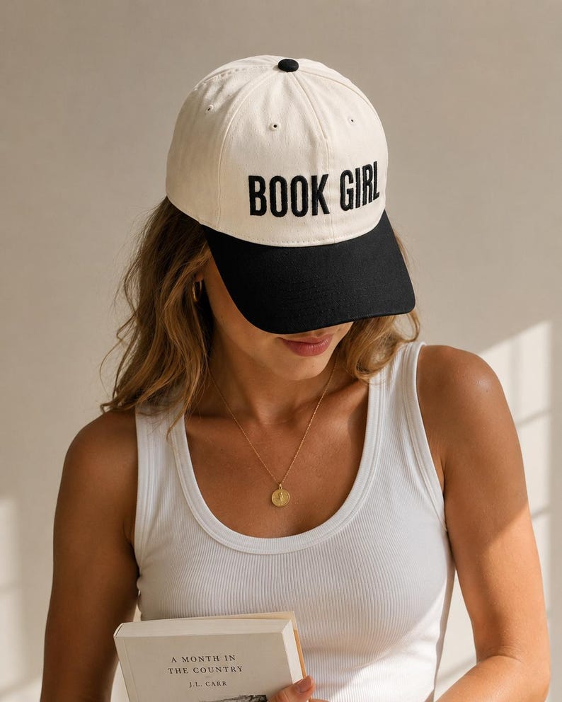 May include: A cream-colored baseball cap with a black brim and the embroidered text "BOOK GIRL" in black. The cap has a black button on top.