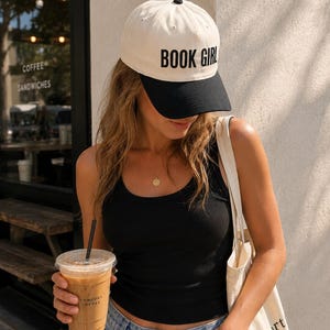 May include: A cream-colored baseball cap with a black brim and the text "BOOK GIRL" in black. The cap is worn by a person wearing a black tank top and blue jeans. The person is holding a coffee drink in a clear plastic cup.