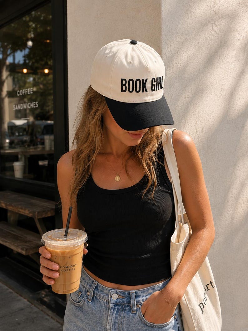 May include: A cream-colored baseball cap with a black brim and the text "BOOK GIRL" in black. The cap is worn by a person wearing a black tank top and blue jeans. The person is holding a coffee drink in a clear plastic cup.